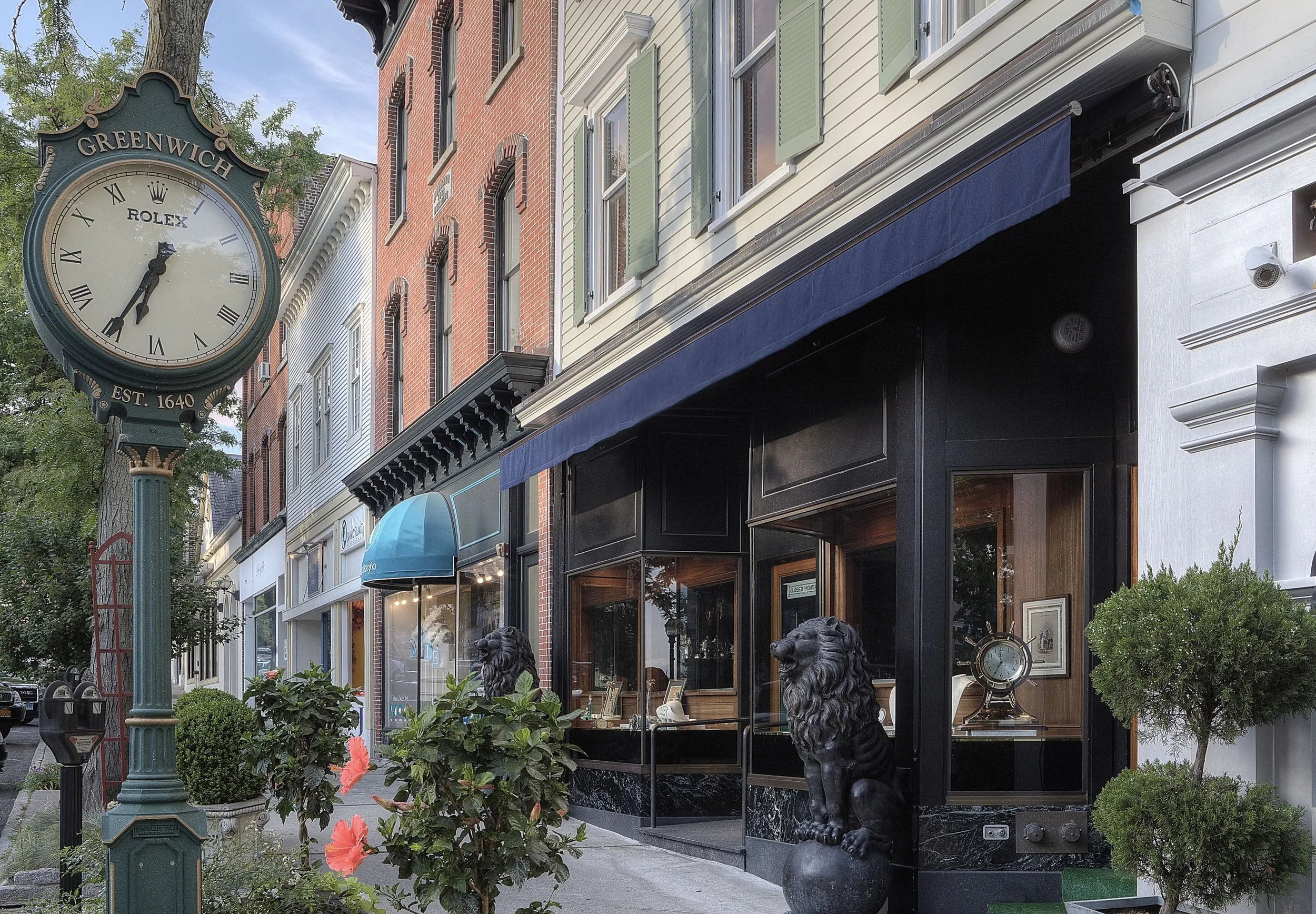 Historic Greenwich Avenue storefronts with a green Rolex street clock and leafy planters in the foreground