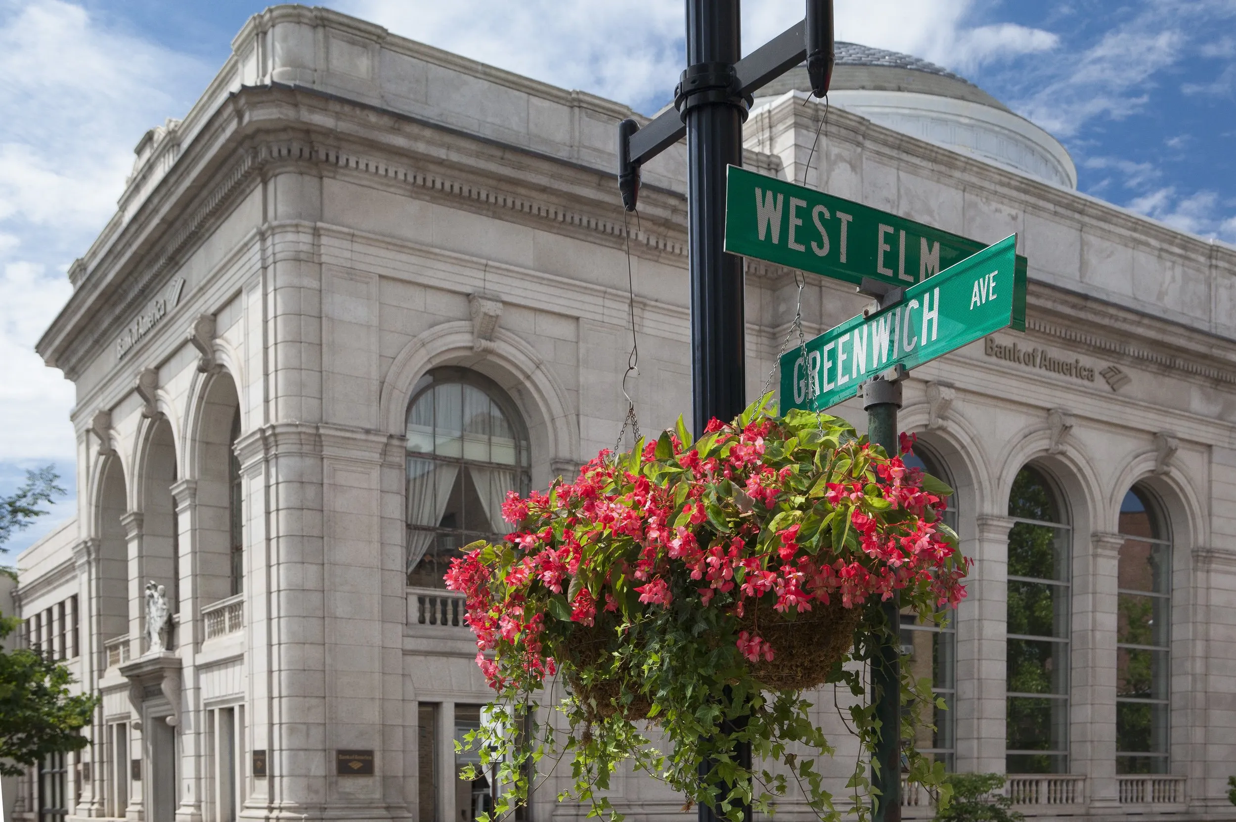 Street-sign post at West Elm St. and Greenwich Ave. framed by hanging pink flowers and a classical stone bank