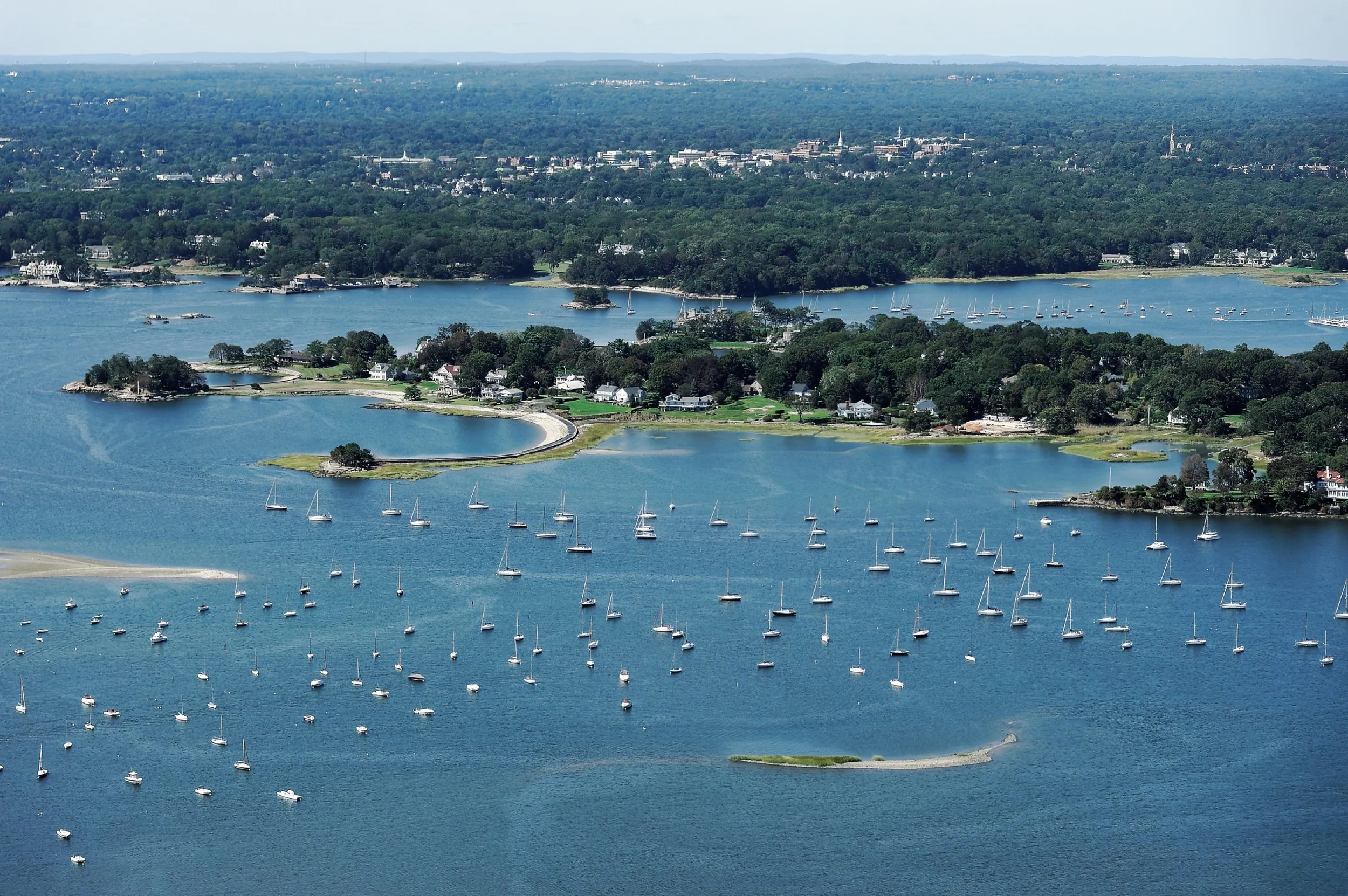Aerial panorama of Greenwich Harbor dotted with moored sailboats, wooded peninsulas and distant shoreline