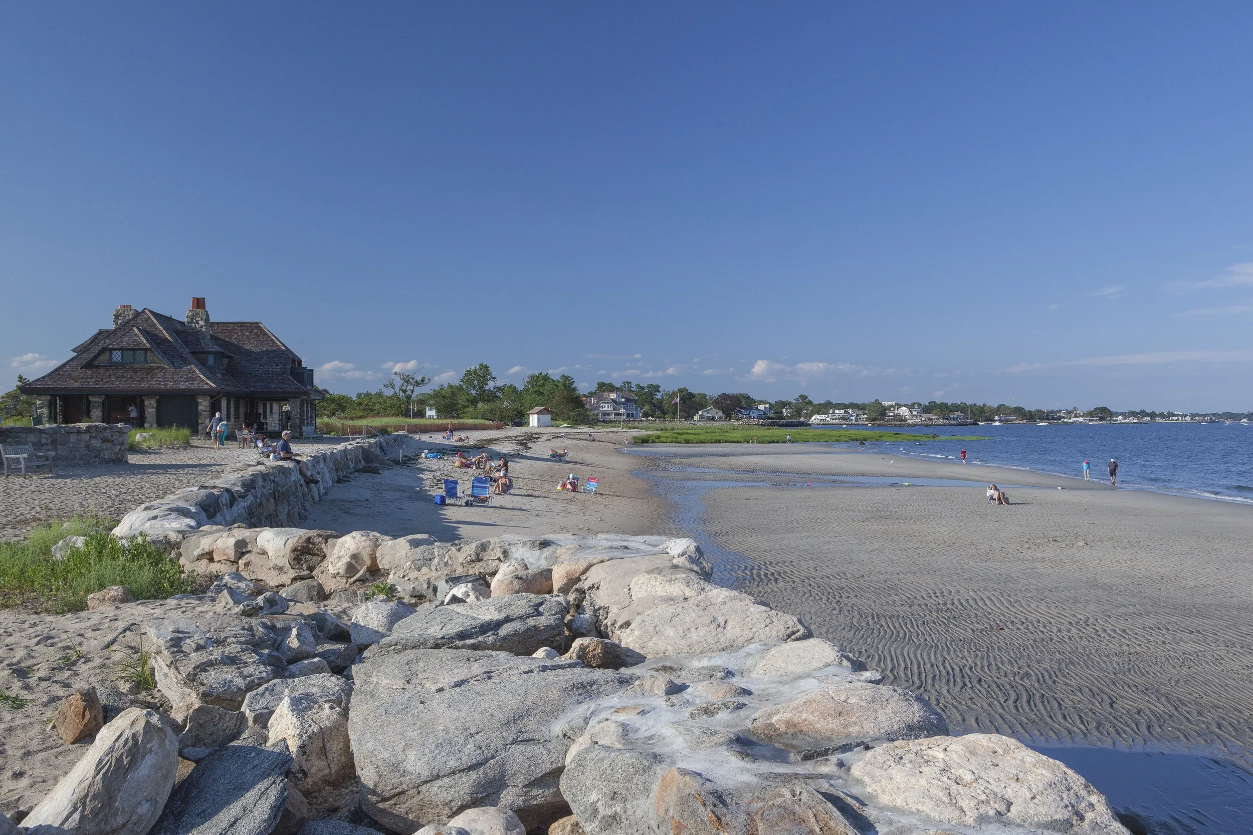 Serene sandy beach with a rocky jetty, shingle-style cottage and families relaxing along Long Island Sound